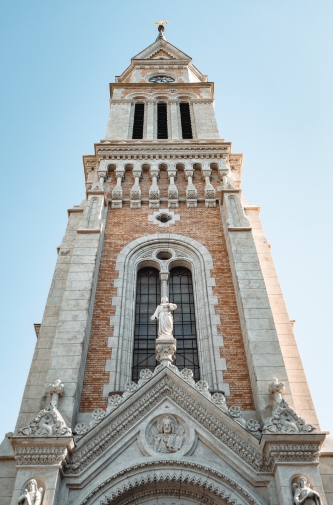 A kép egy impozáns templomtornyot ábrázol, amely a neogótikus és neoromán építészeti stílusok keverékét mutatja. A torony téglából és kőből épült, és több szintre tagolódik. Az alsó részen egy nagy, íves ablak látható, amelyet kőkeretezés vesz körül. Az ablak felett egy fehér kőszobor áll, valószínűleg egy szentet vagy Jézust ábrázolja. A torony középső része téglából készült, díszítő kőelemekkel. Ezen a részen egy sor kis boltíves nyílás látható, amelyek valószínűleg dekoratív célokat szolgálnak. A torony felső részén három nagy, keskeny ablak található, felettük pedig egy kerek óra. A torony csúcsán egy kereszt látható. Az egész szerkezet részletgazdag díszítésekkel van ellátva, beleértve a faragott kőmunkákat, szobrocskákat és geometriai mintákat. A torony a kék ég előtt magasodik, ami kiemeli annak impozáns megjelenését és vertikális vonalait. A kép alulról felfelé készült, hangsúlyozva a torony magasságát és monumentalitását, ami egy lenyűgöző látványt nyújt a szemlélőnek. (Kiemelt képünk illusztráció, forrás: a templom weboldala; a kép leírását a Claude generálta)