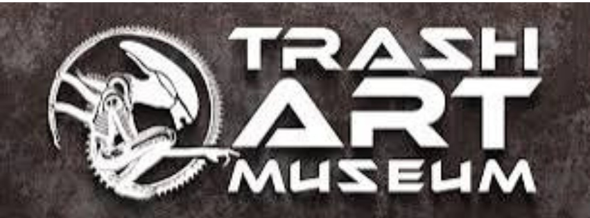 A kép egy logót ábrázol, amelyen a "TRASH ART MUSEUM" felirat látható. A logó bal oldalán egy stilizált, kör alakú embléma található, amely egy xenomorf-szerű alien fejet ábrázol (hasonlót az Alien filmekből ismerthez), oldalnézetből. A szöveg és a logó fehér színű, míg a háttér sötét, texturált szürke. A betűtípus modern, szögletes kialakítású, ami futurisztikus hatást kelt. Az egész design ipari, sci-fi jellegű megjelenést sugall.