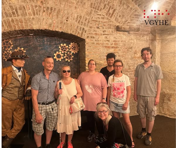A képen nyolc ember látható, egy boltíves, téglás fal előtt. Az emberek különböző korúak és öltözékűek. Az egyik személy steampunk stílusú öltözetben van, kalapban és szemüvegben. Néhányan napszemüveget viselnek, és különböző pózokban állnak vagy ülnek. Hátul fogaskerekeket ábrázoló dekoráció található.