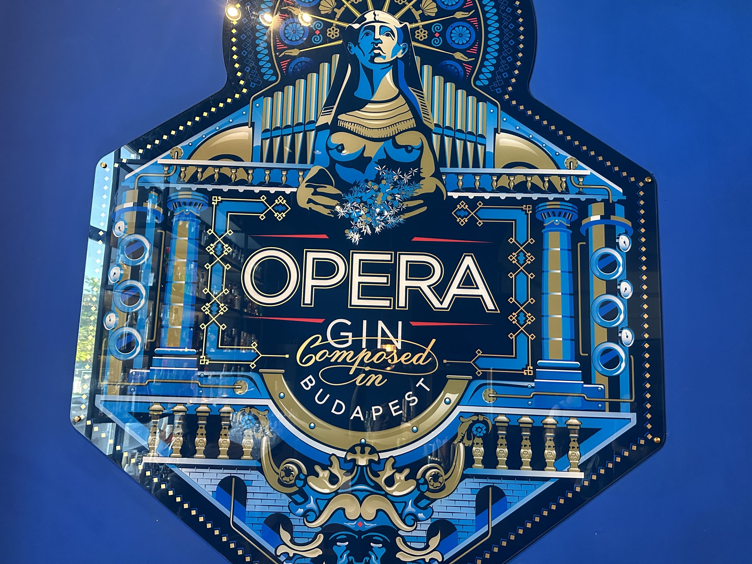 Az Opera Gin díszes, kék, arany és fekete címkéje. Középen az „OPERA GIN Composed in Budapest” felirat látható, fölötte egy stilizált, egyiptomi ihletésű női alak, körülötte pedig zenei és építészeti motívumok, amelyek a budapesti Operára utalnak.